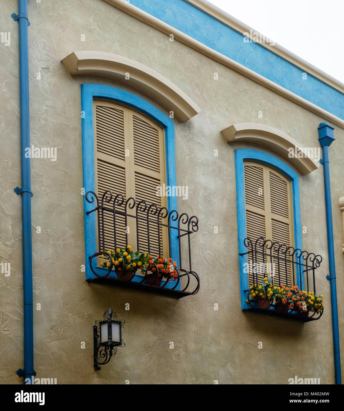 Two shuttered windows with wrought iron window boxes, bright flowers & blue frames. Two blue drain pipes. Retro light on building under one window. Stock Photo