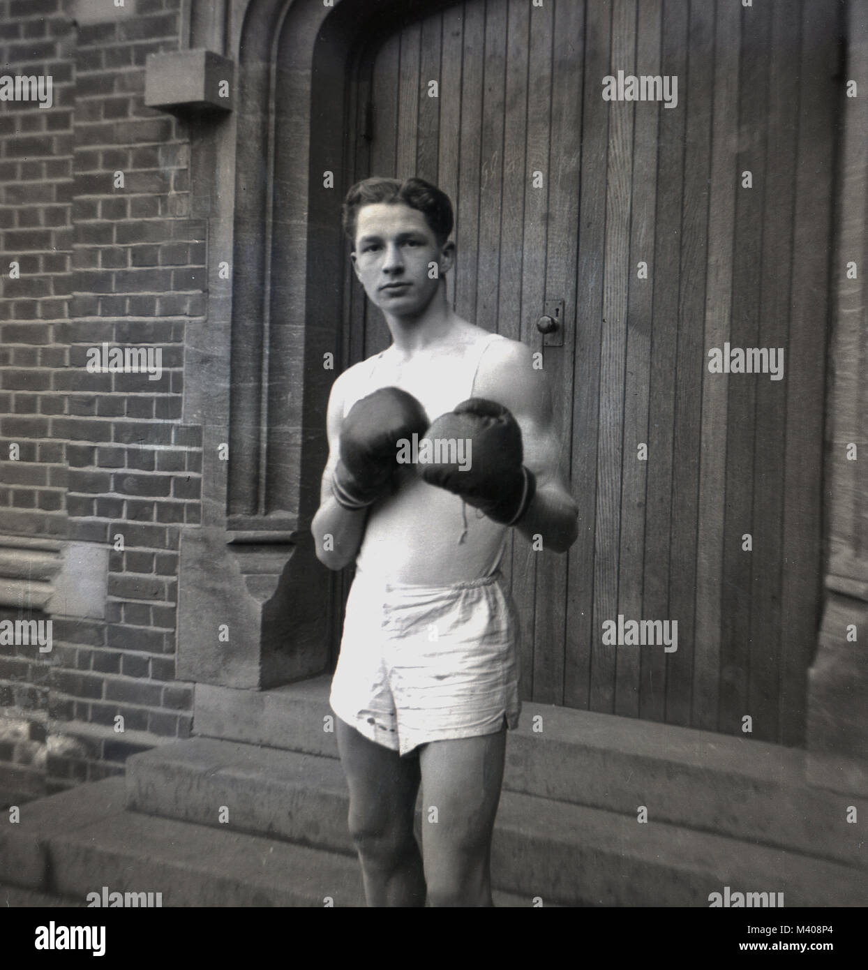 1950, historical...the 'noble art'...a young male school pupil standing outside a large wooden doorway at Bancrofts School, Woodford Green, England, UK, wearing a sports vest and shorts and boxing gloves and making a punching pose or stance. Boxing was a sport taught in both British state and public (private) schools at this time, and apart from the health and fitness benefits, pupils learnt that fighting took place in a ring, not on a street. Stock Photo