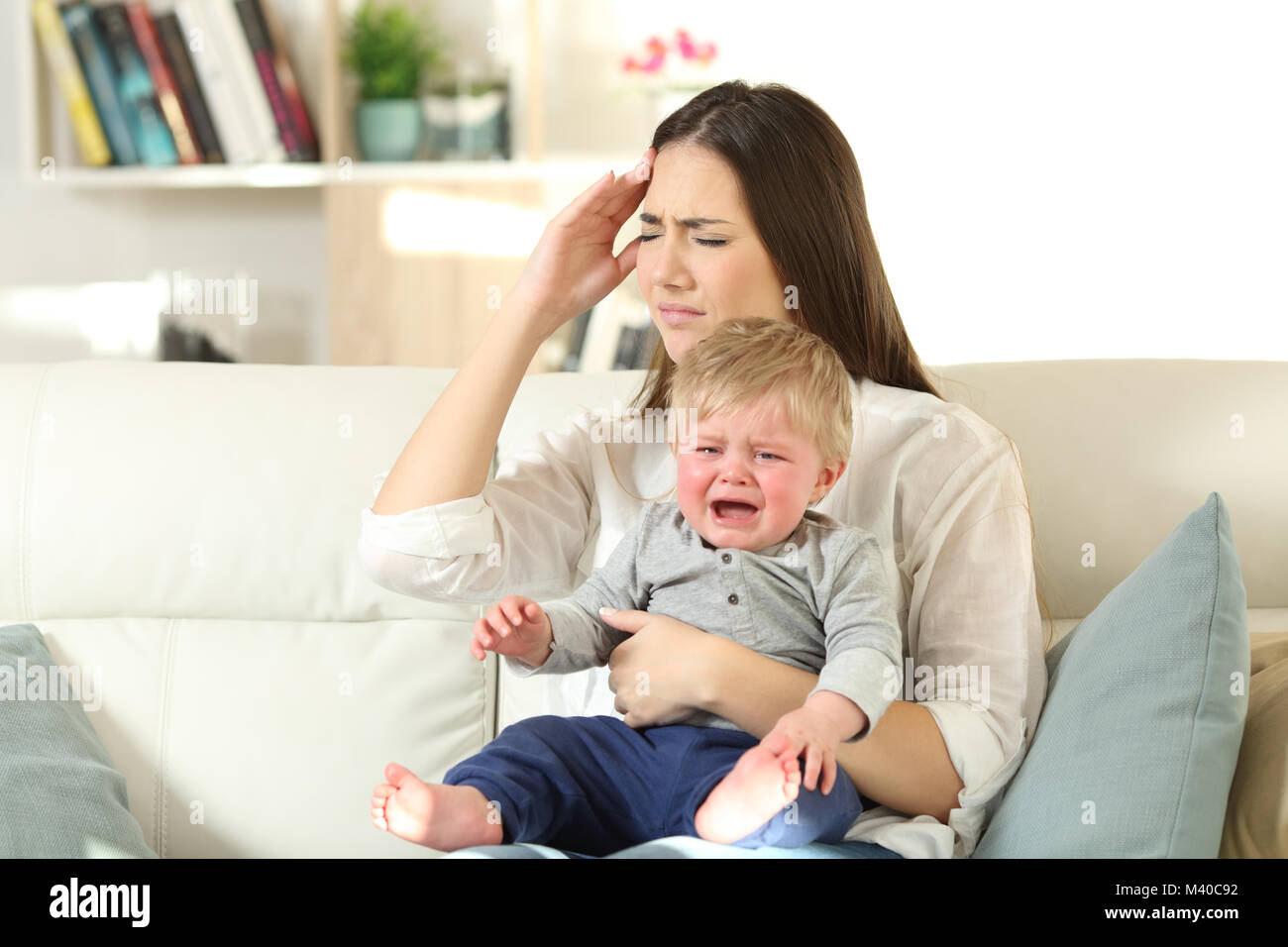 Mother suffering and baby crying desperately sitting on a couch in the living room at home Stock Photo