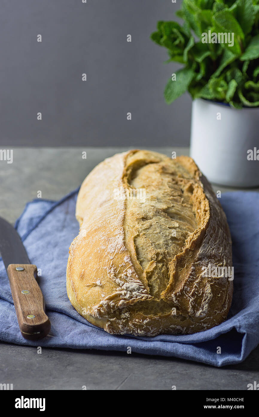Freshly Baked Hand Crafted Rustic Bread Loaf on Blue  Linen Towel on Dark Kitchen Table Knife Green Herbs. Natural Light Authentic Style. Lifestyle Ph Stock Photo