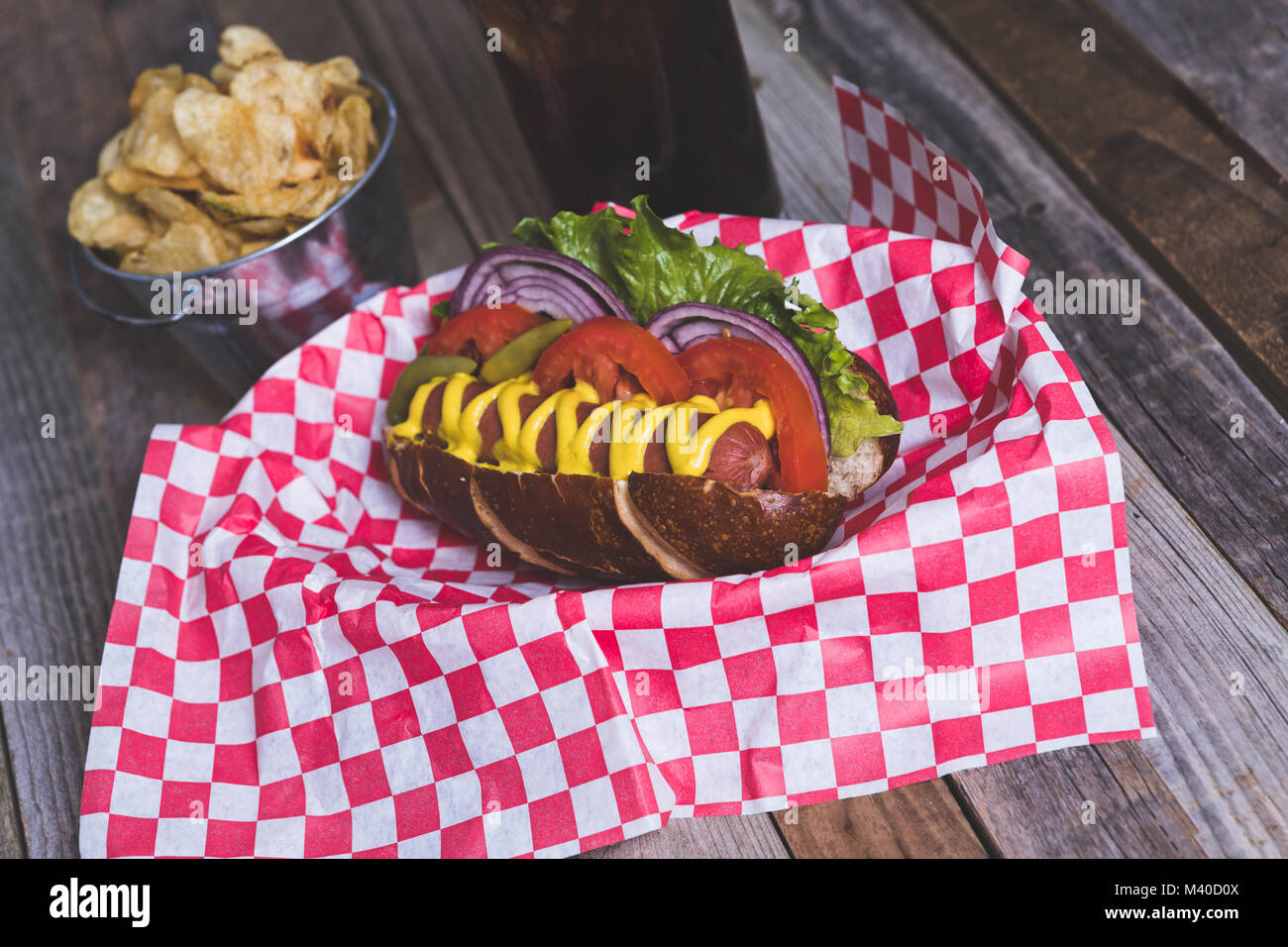 Gourmet hot dog with chips and drink in a basket with gingham napkin. Tabletop, side view. Stock Photo