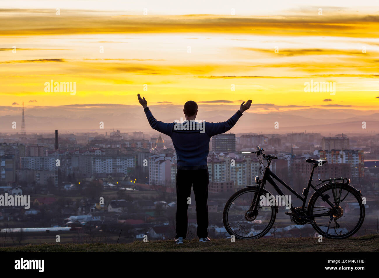 Dark silhouette of man standing near a sports bicycle with the night city view behind. Active life and travel concept. Sunset over Ivano Frankivsk cit Stock Photo