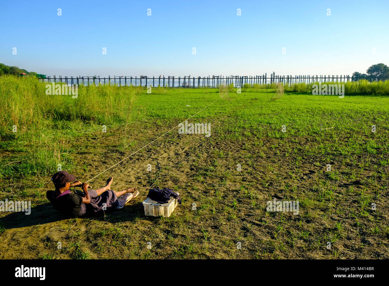 A boy is catching singing birds with a trap on a meadow next to U Bein Bridge Stock Photo