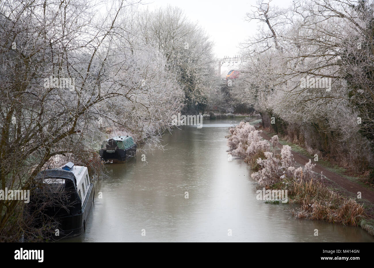 Narrowboats on the Grand Union Canal at Linslade, Bedfordshire, with hoar frost on nearby trees and a red train in the background. Stock Photo