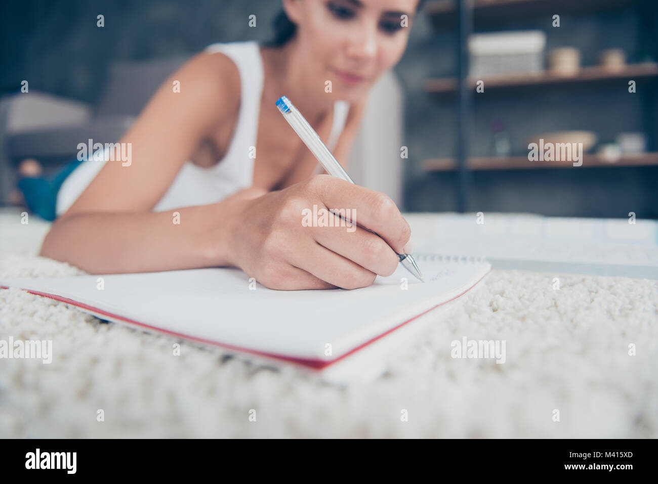 Close up portrait of woman hand with pencil writing in copybook, blurred background of girl laying on stomach on carpet Stock Photo