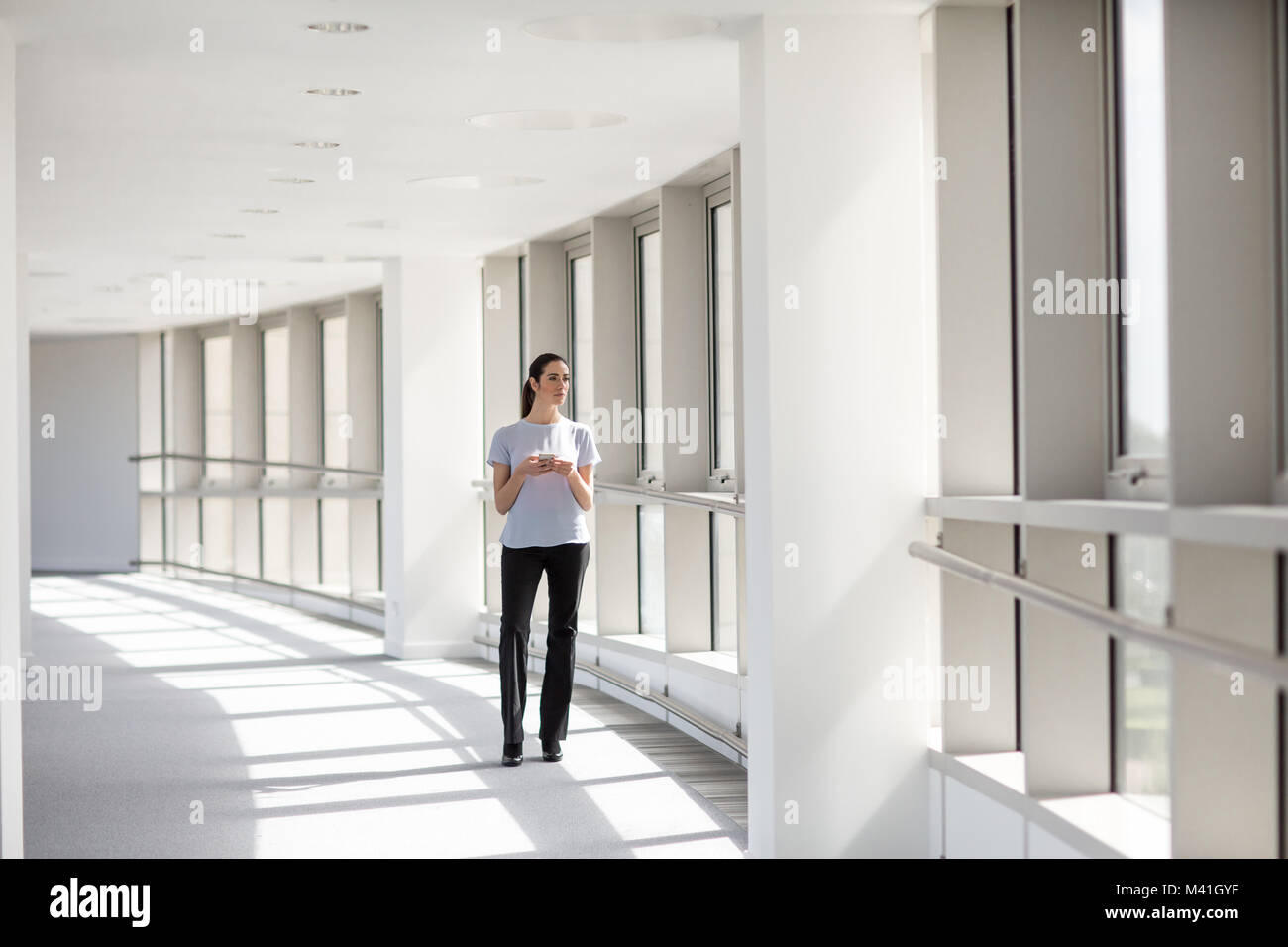 Businesswoman holding smartphone looking out of window in office Stock Photo