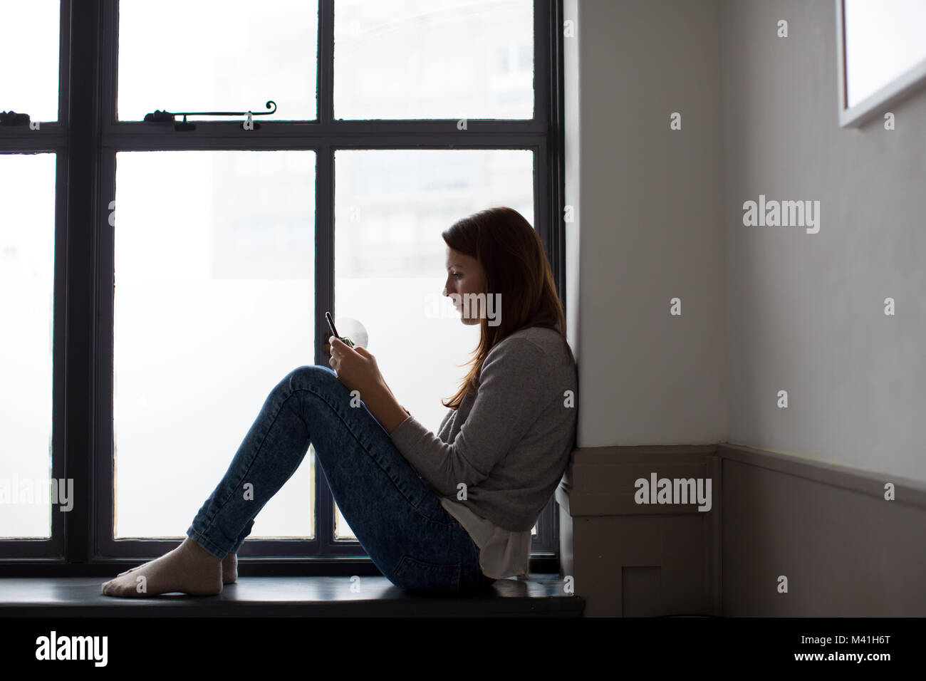 Young adult female sitting on window ledge with smartphone Stock Photo