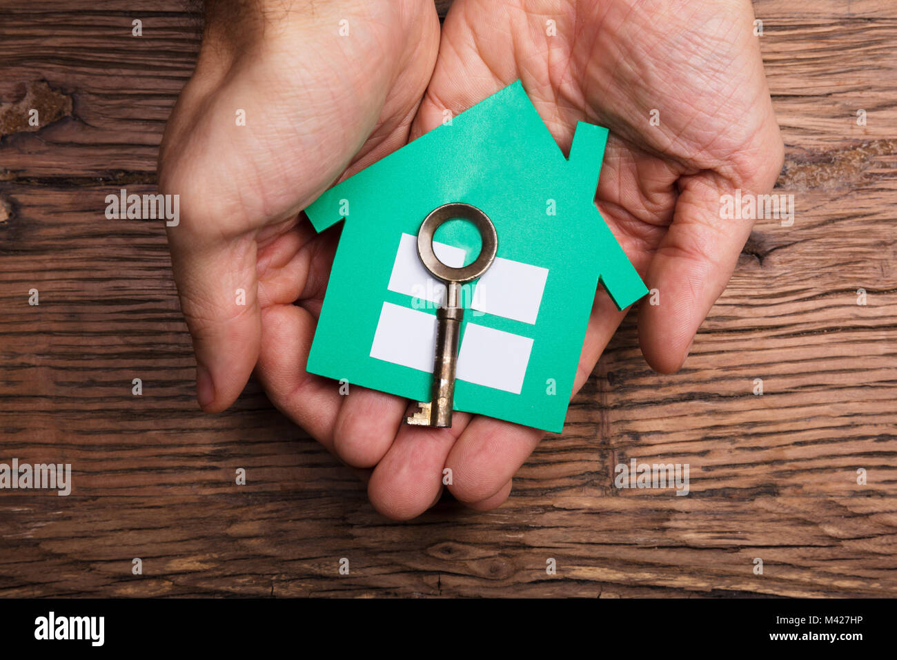 High Angle View Of Hands Holding Paper House With House Key Against Wooden Table Stock Photo