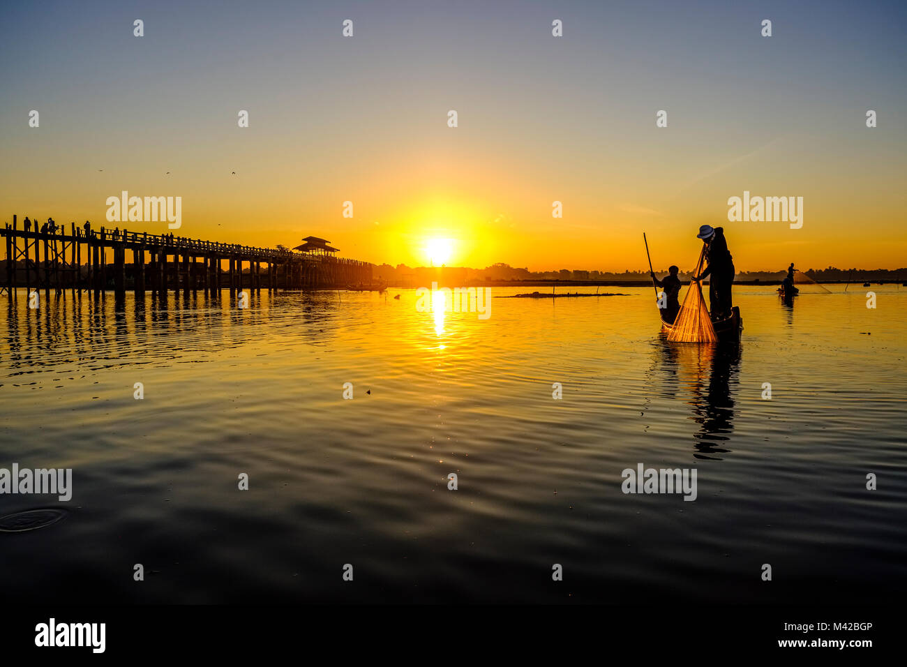 A fisherman on a boat is pulling his net out of Taungthaman Lake next to U Bein Bridge at sunrise Stock Photo