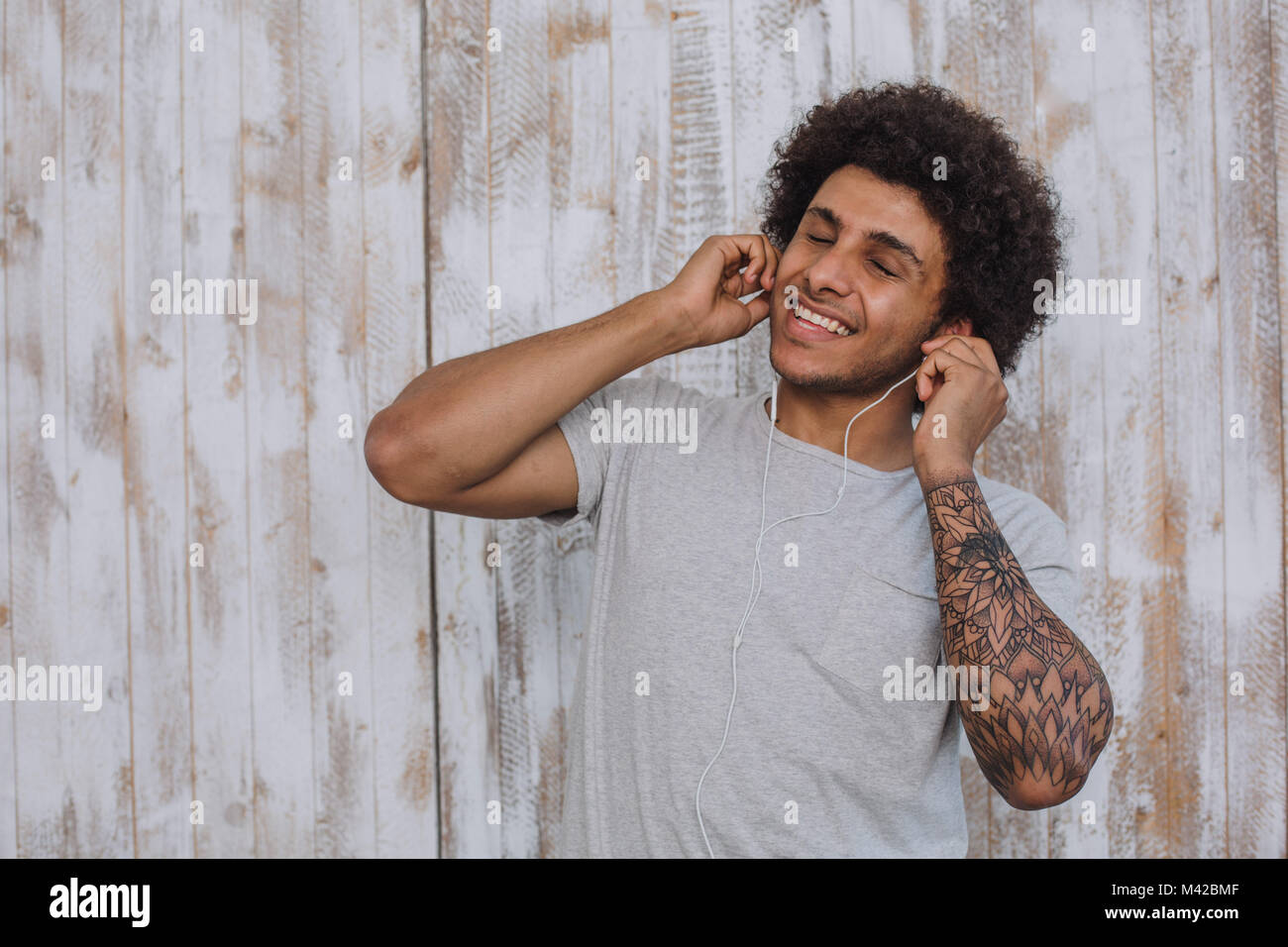 enjoy the sound. Mulatto man, with curly hair wearing headphones, closed his eyes and smiling against retro wooden wall Stock Photo