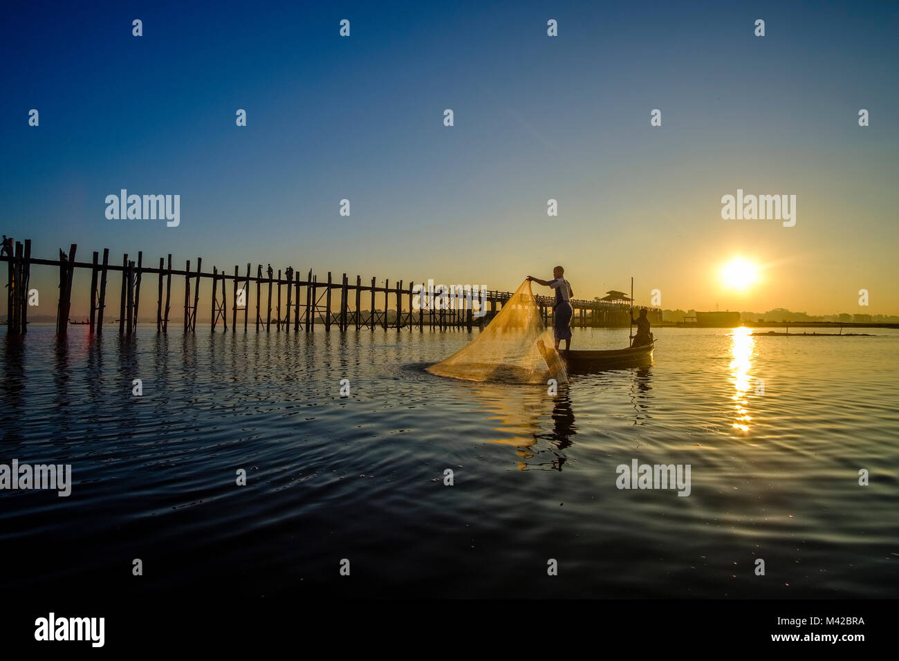 A fisherman on a boat is pulling his net out of Taungthaman Lake next to U Bein Bridge at sunrise Stock Photo