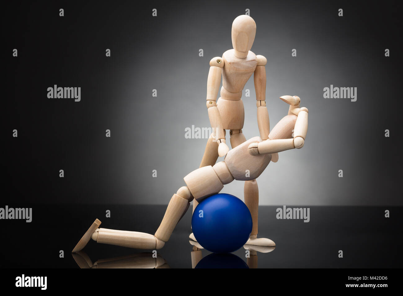 Trainer Assisting Wooden Figurine While Exercising On Blue Fitness Ball Against Grey Background Stock Photo