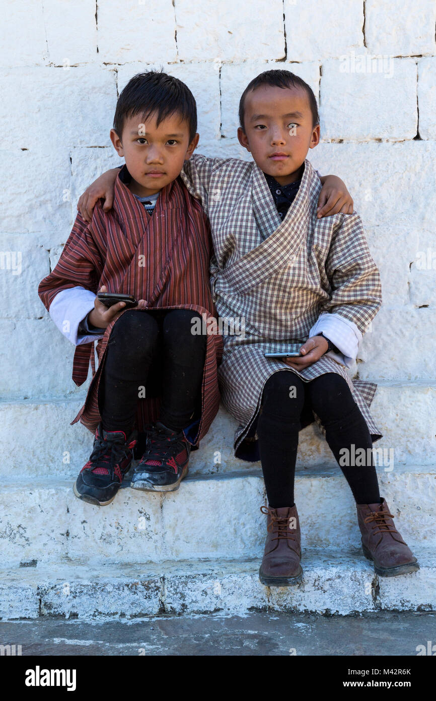 Punakha, Bhutan.  Two Young Boys Wearing Traditional Gho, with Cell Phones, Chimi Village. Stock Photo