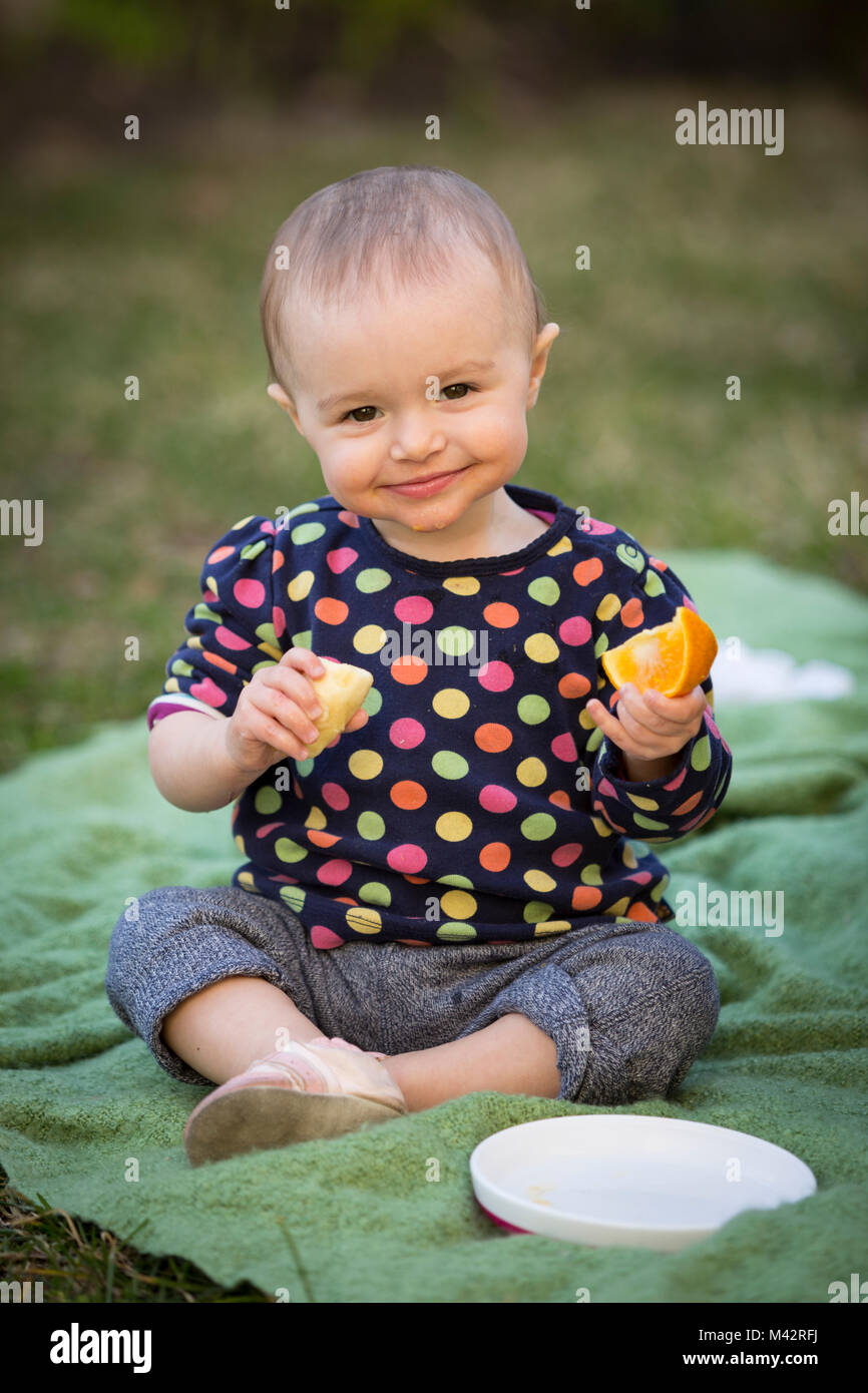 A young toddle enjoys a fruit snack of orange slices and bananas in a back yard garden. Stock Photo