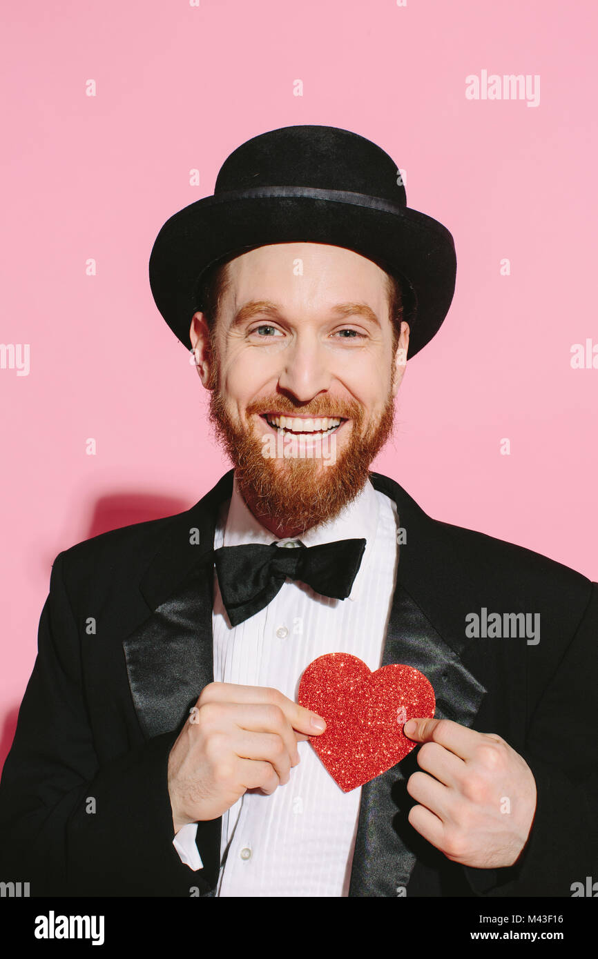 smiling man in tux and top hat holding a red heart on his chest Stock Photo