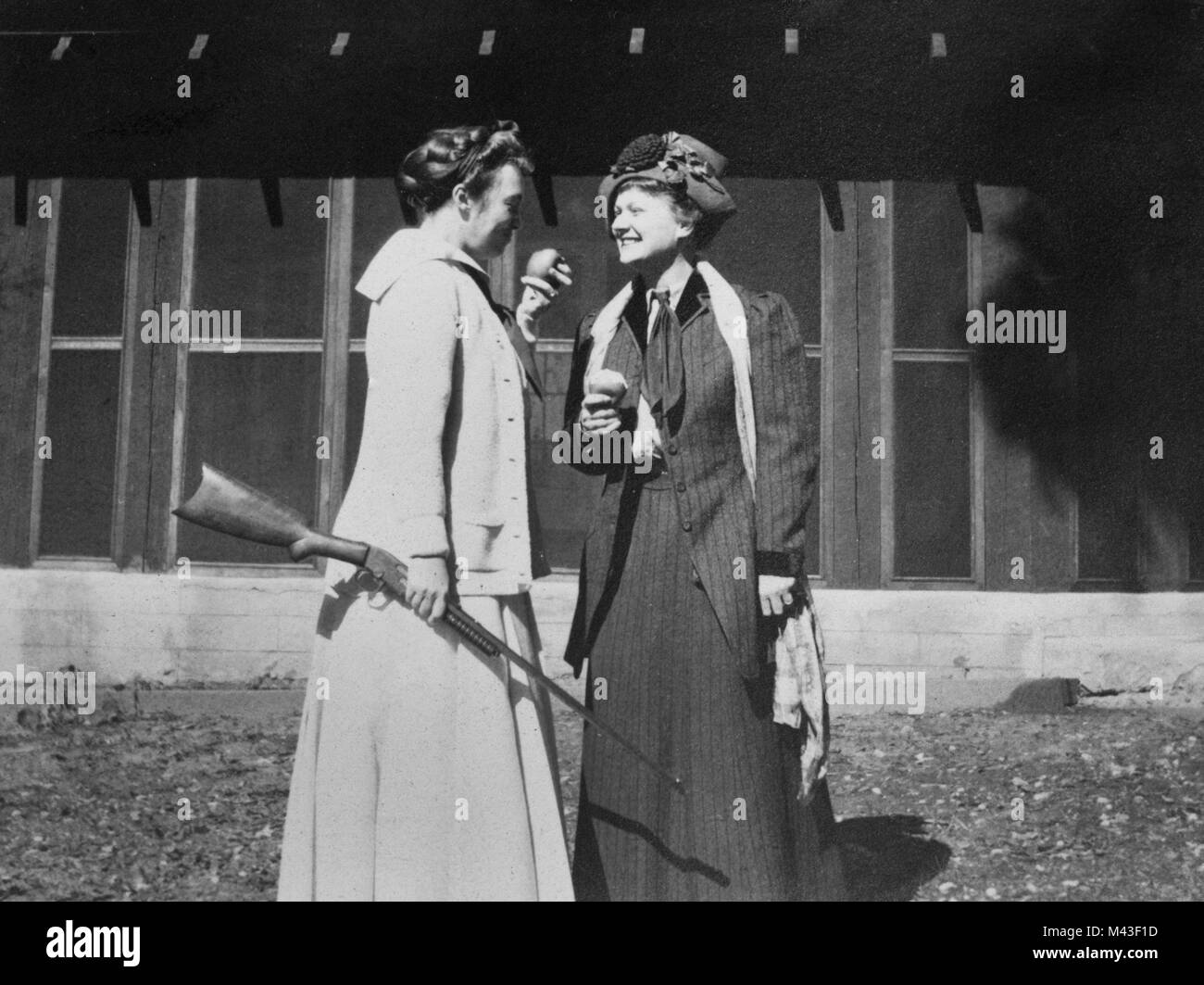 A woman holds a rifle and eats an apple while talking with another woman, ca. 1910. Stock Photo