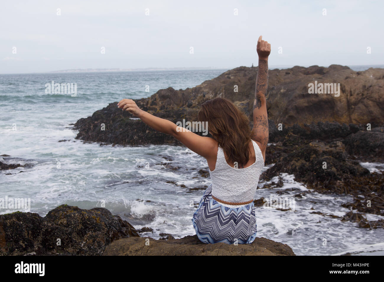 Back view of happy tattooed woman sitting on rocks by the ocean with her arms raised Stock Photo