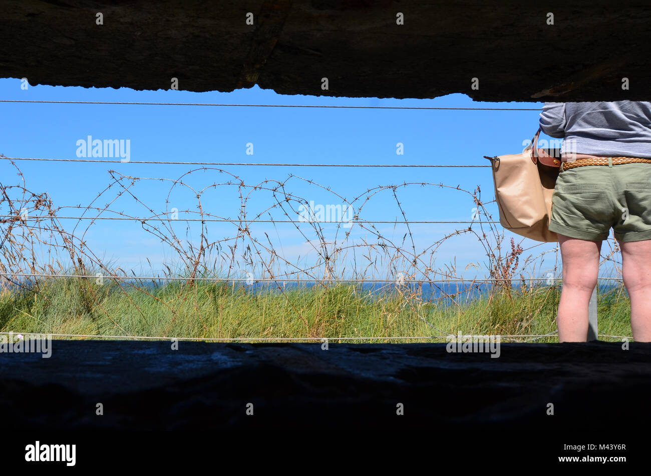 View out from inside German defensive bunker on cliff top at Omaha Beach, Normandy to barded wire and tourist looking out to D-Day landing site Stock Photo