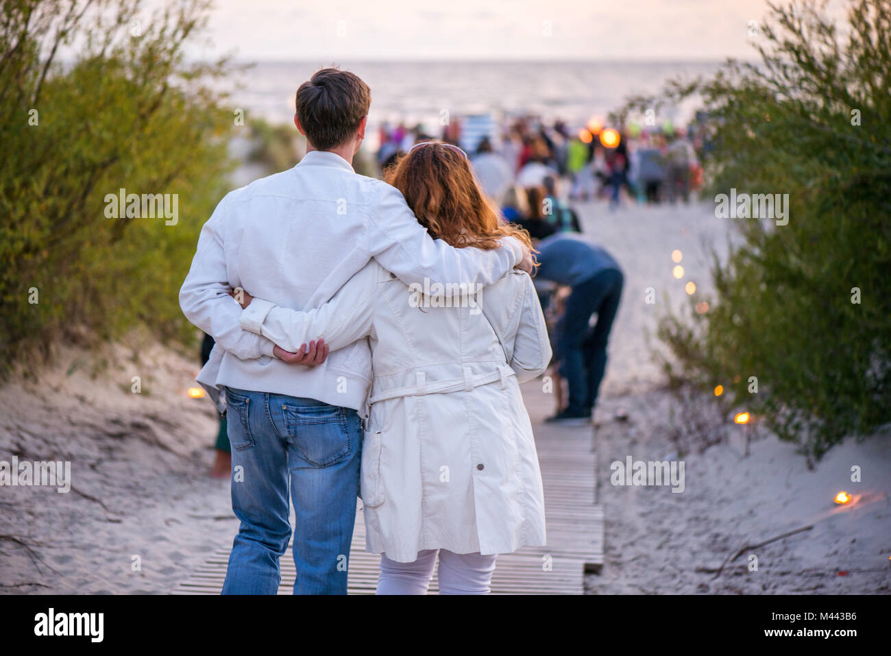 Happy couple hugging and walking on seaside on lit candle path during sunset. Romantic summer evening, love is in the air. Stock Photo