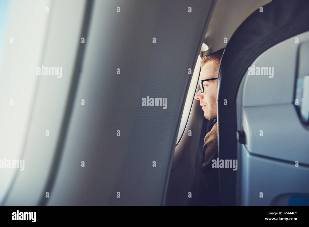 Comfortable traveling by airplane. Young man (traveler) with eyeglasses looking from window during flight. Stock Photo