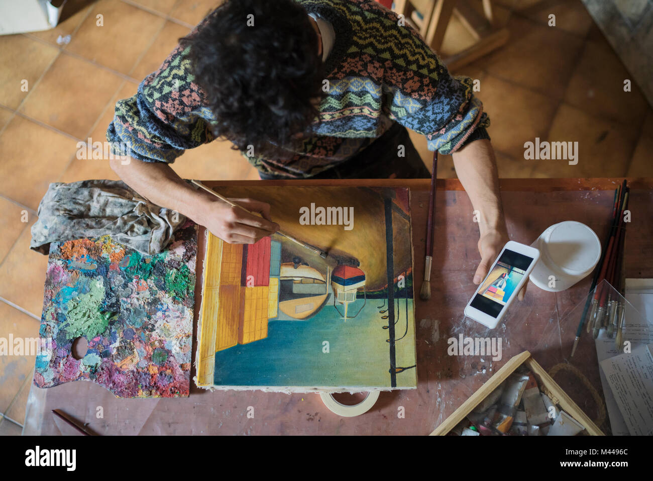 Male artist looking at smartphone while painting canvas in artists studio, overhead view Stock Photo