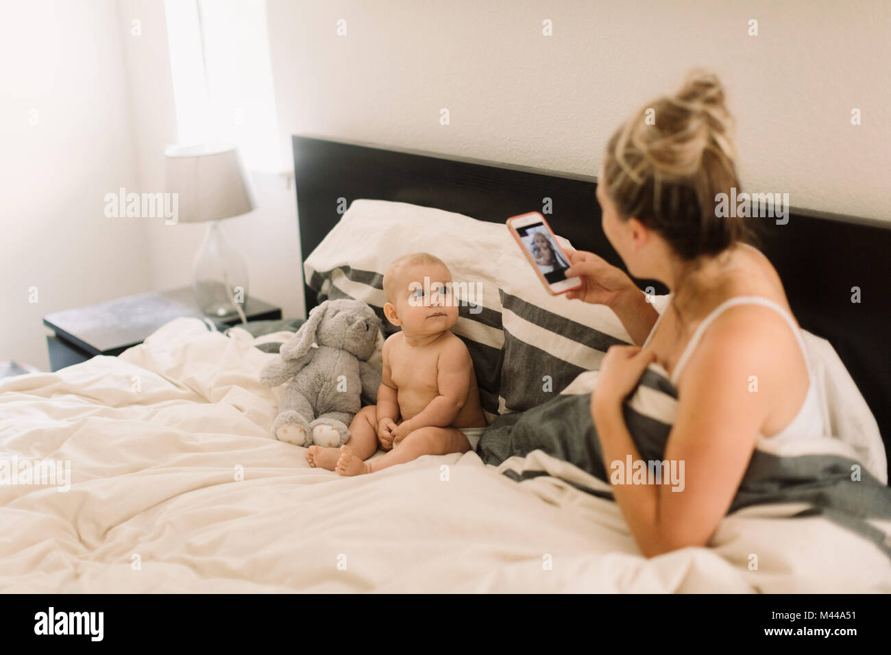 Mother taking smartphone photograph of baby daughter sitting up in bed with soft toy Stock Photo