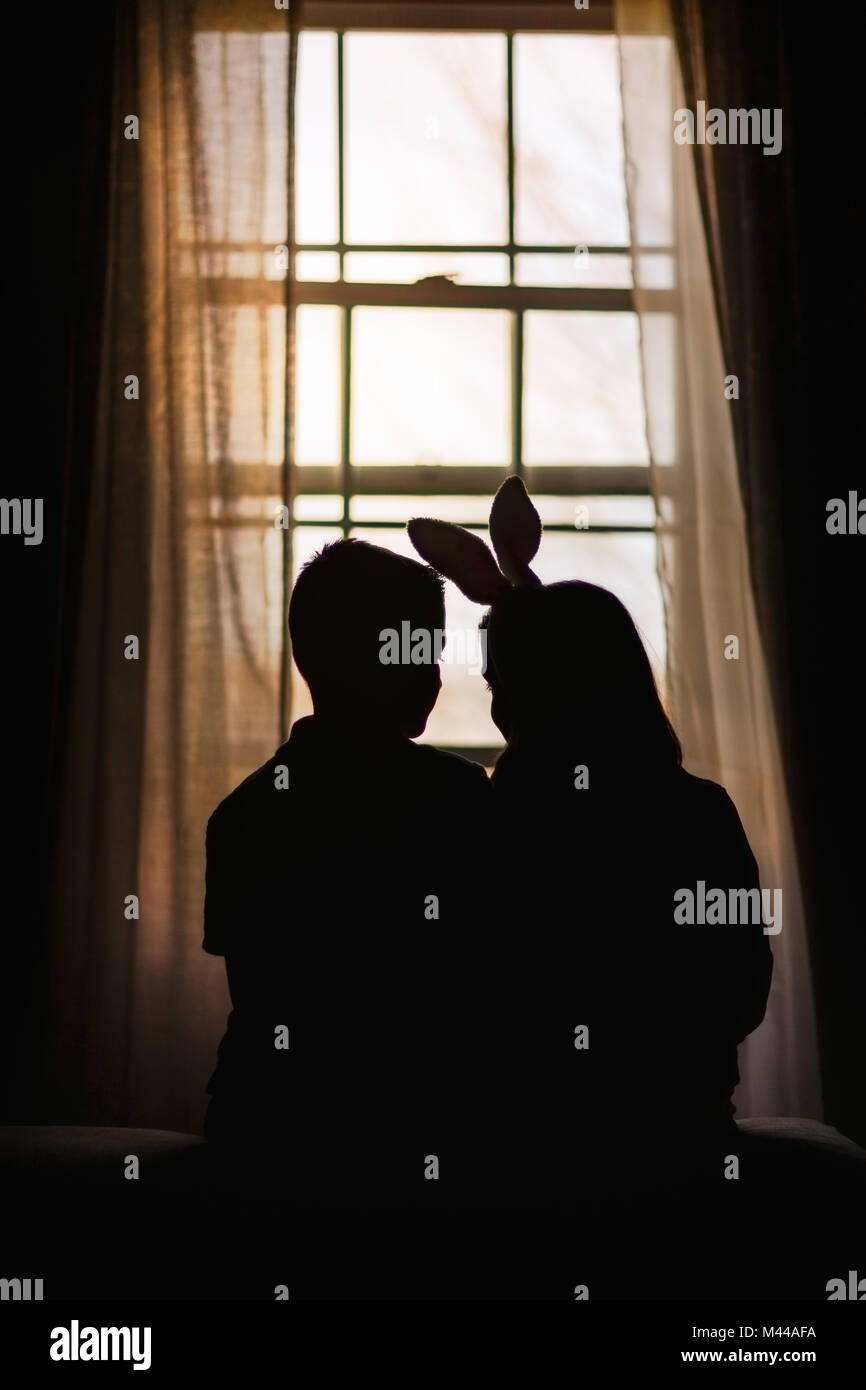 Silhouette of boy and girl, sitting in front of window, girl wearing bunny ears, rear view Stock Photo