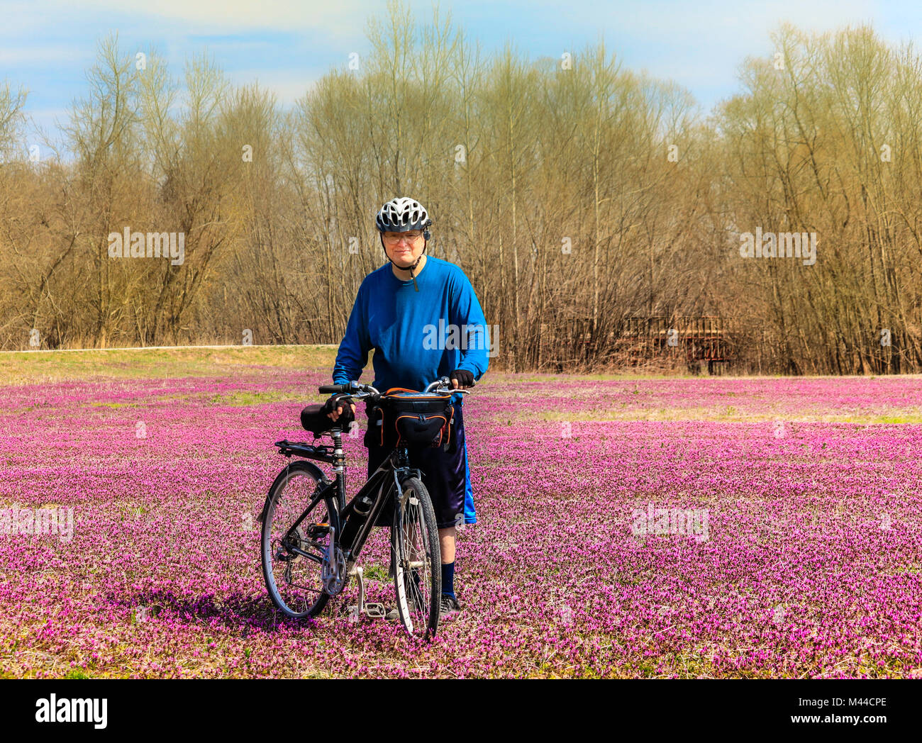 Senior bicyclist stands in the field of wildflowers smiling; spring the Midwest; trees in the background Stock Photo