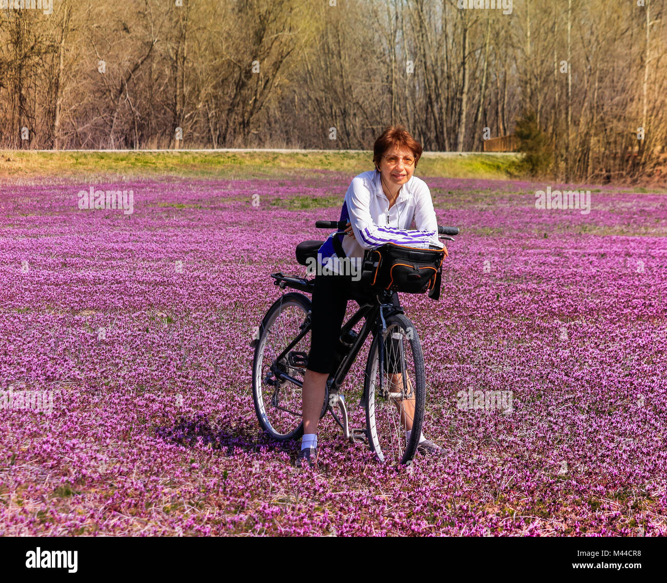 Older female bicyclist stands in the field of wildflowers leaning on her bicycle and smiling; spring in Missouri, Midwest Stock Photo