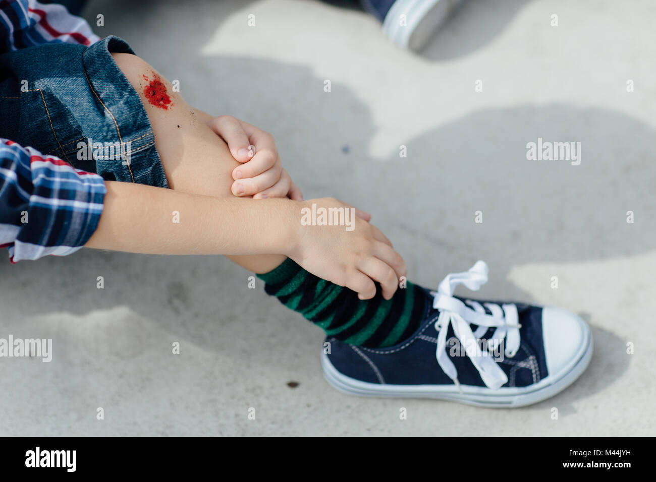 Injured boy sitting on concrete floor and  looking at his scraped knee Stock Photo