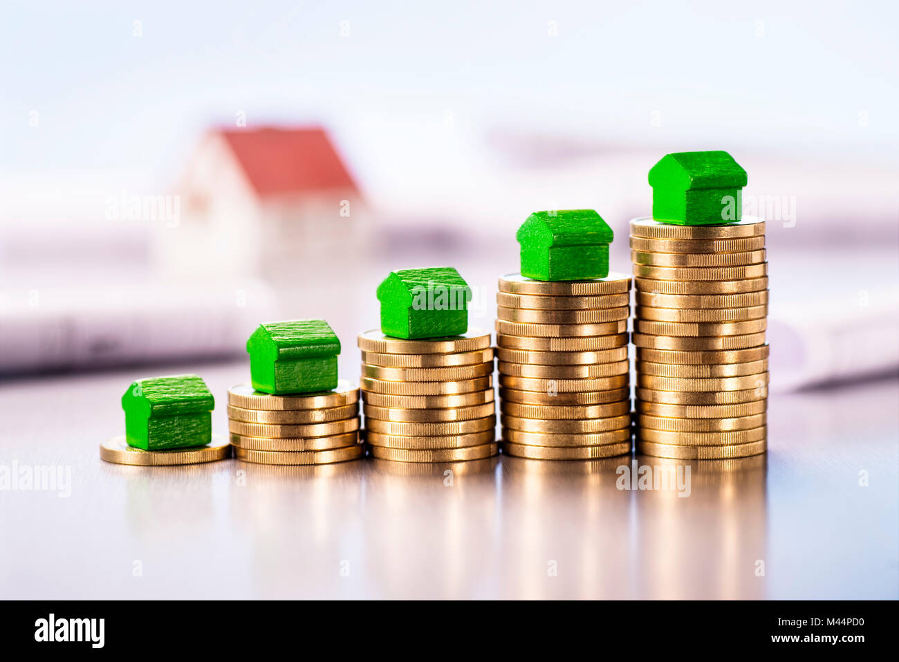 Small green houses standing on stacks of coins with blueprints and architectural model in the background. Stock Photo