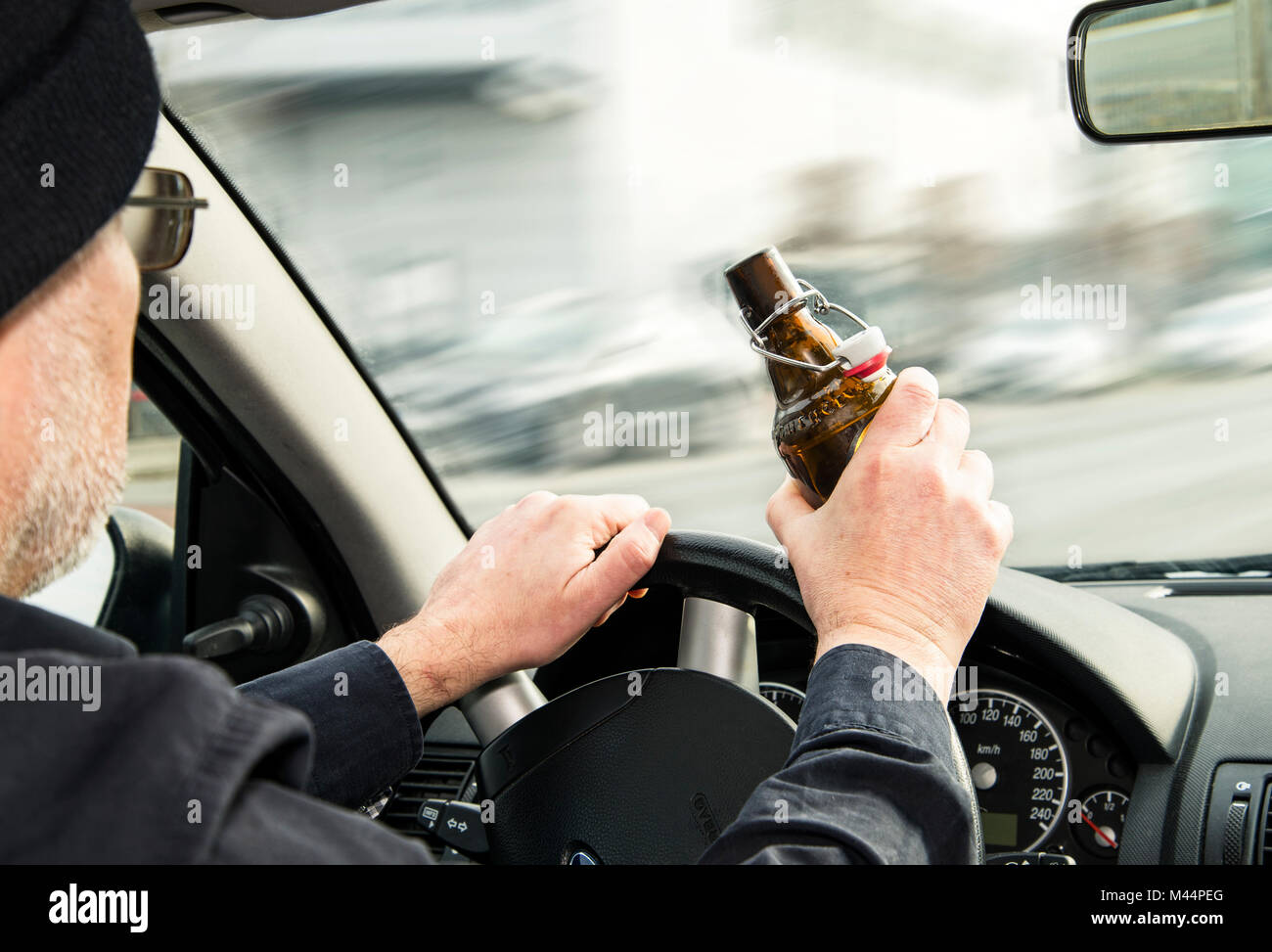 Man holds an opened bottle of beer in one hand and covers the steering wheel of a car with the other. Stock Photo