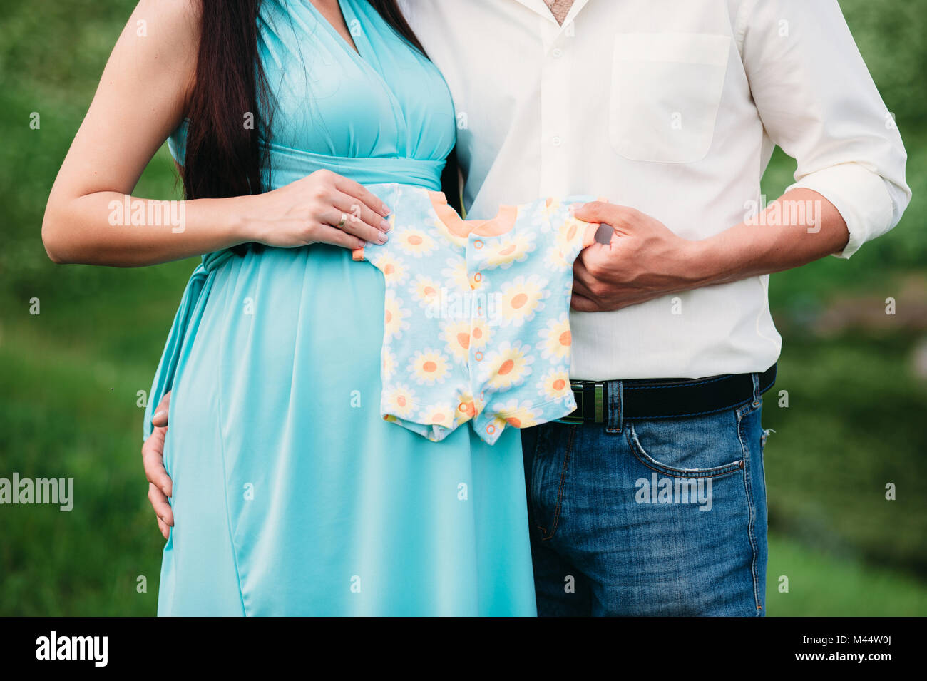 future paremts holding kids clothes near pregnant belly. Family, materniry, pregnancy concept. Closeup. Stock Photo