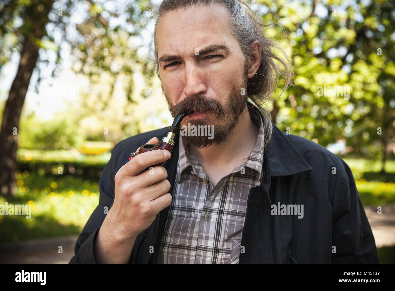 Young serious bearded man smoking pipe in summer park, close-up portrait Stock Photo