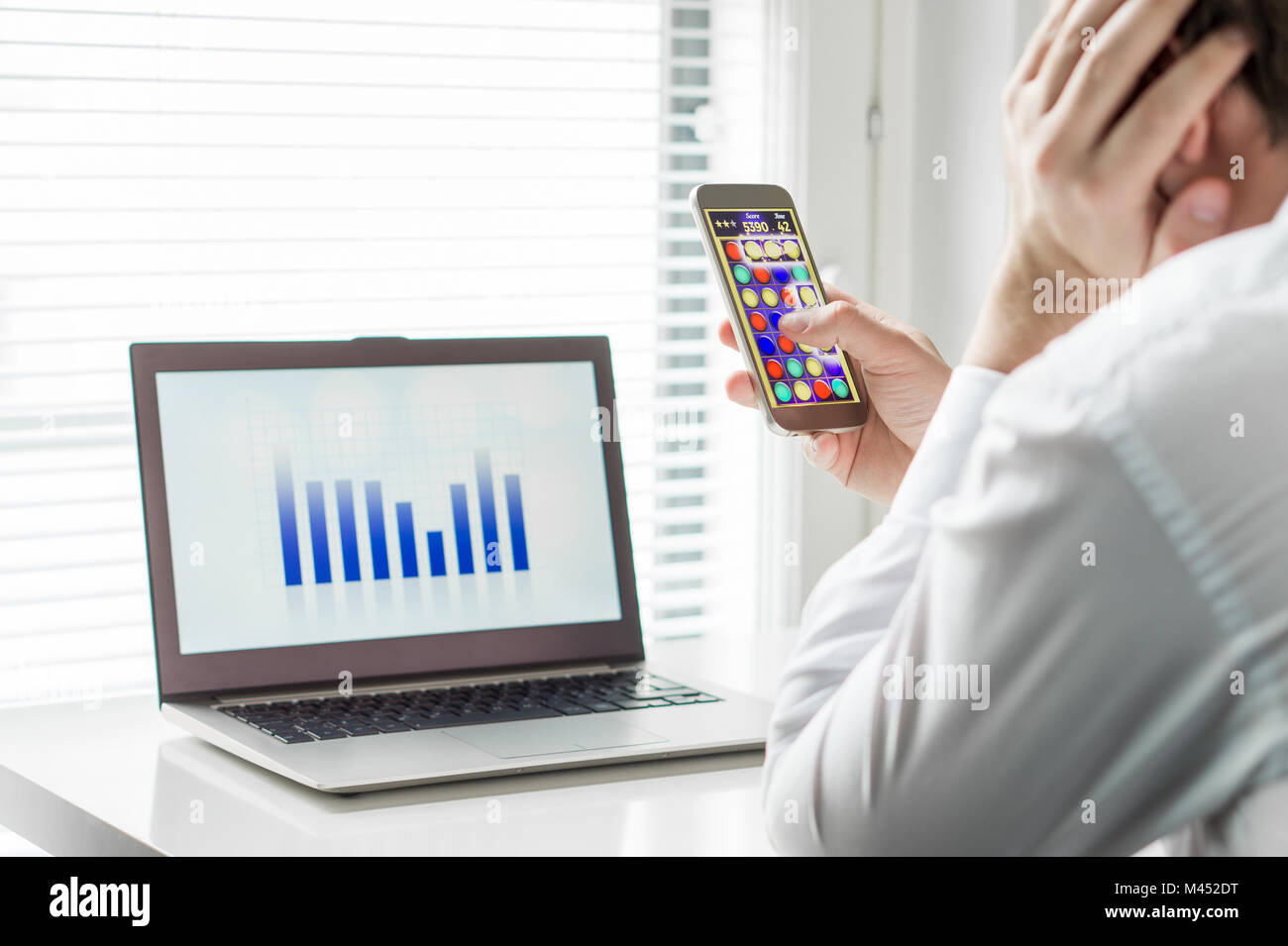 Lazy office worker playing mobile game with smartphone during work hours. Avoiding his job and leaning head against hand. Bored employee. Stock Photo