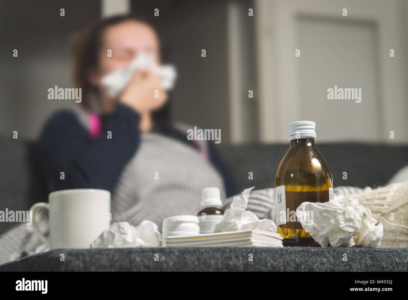 Sick woman sneezing to tissue. Medicine, hot beverage and dirty paper towels in front. Girl caught cold. Cough syrup and handkerchiefs on table. Stock Photo