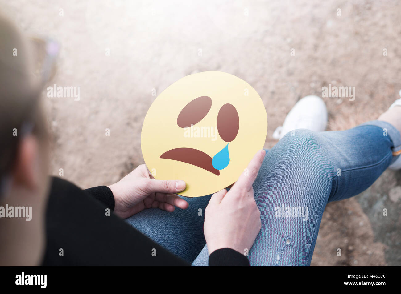 Crying paper emoticon in hand. Depressed woman holding printed sad smiley face and sitting on a rock. Modern communication and smiley icon. Stock Photo