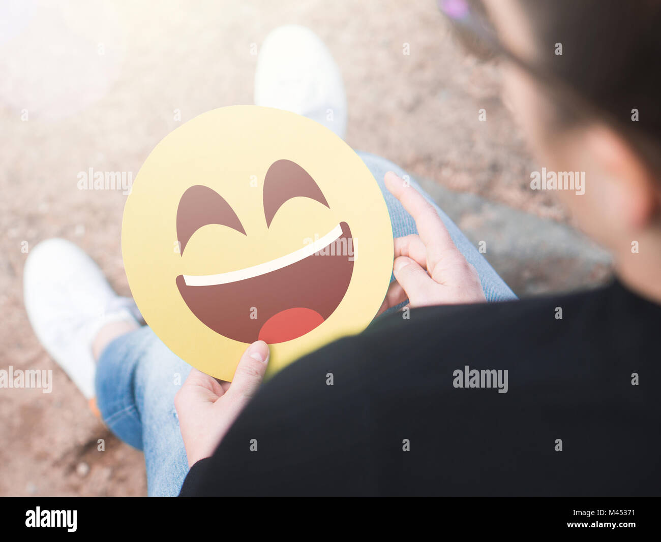 Laughing happy emoticon in hand. Woman holding a cheerful and joyful printed paper smiley face. Happy communication and smiley icon on cardboard. Stock Photo