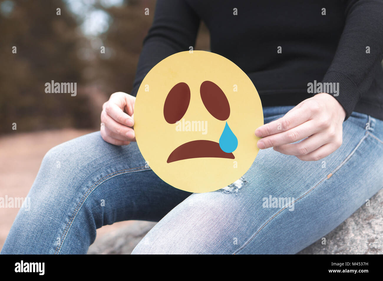 Sad paper emoticon in hand. Depressed woman holding printed crying smiley face and sitting on a rock. Modern communication and smiley icon. Stock Photo