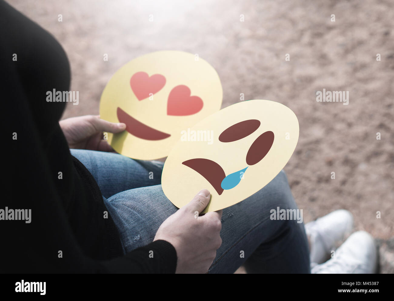 Woman holding 2 cardboard emoticons in hand. Love heart and crying smiley faces. Heartbroken or confused girl having mixed feelings about emotions. Stock Photo