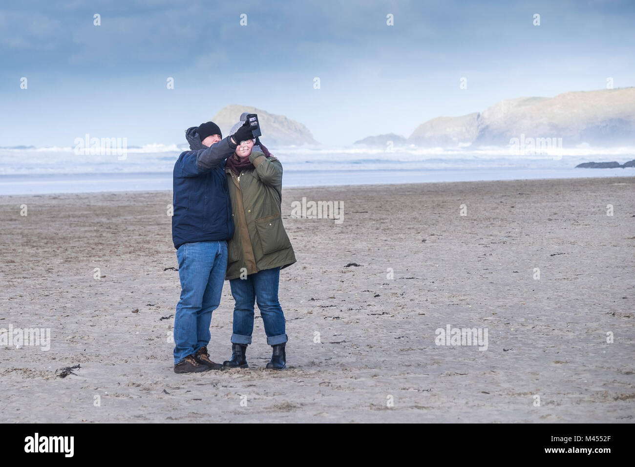 A couple taking a selfie photograph with a smartphone on a windy Perranporth Beach in Cornwall UK. Stock Photo