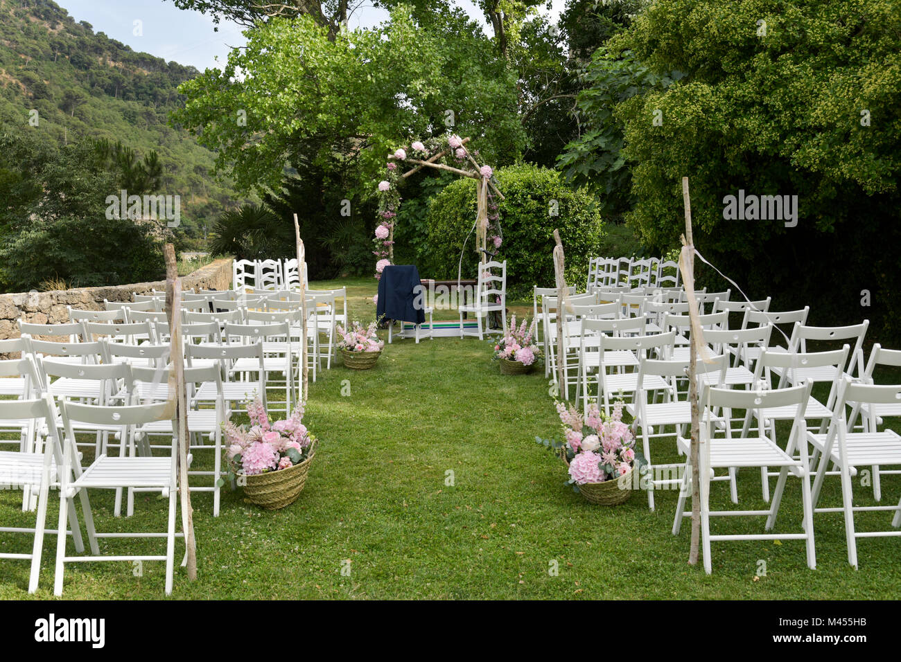 a view of the scene of an open-air wedding ceremony, with some rows of white folding chairs and some arrangements of different pale pink flowers, and  Stock Photo