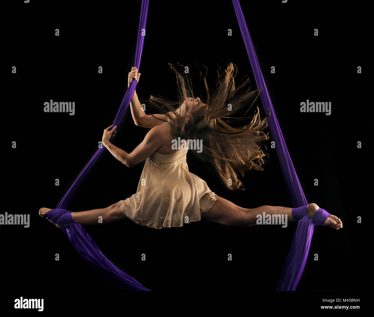 Young female aerial acrobat doing the splits with feet wrapped in silk rope against black background Stock Photo