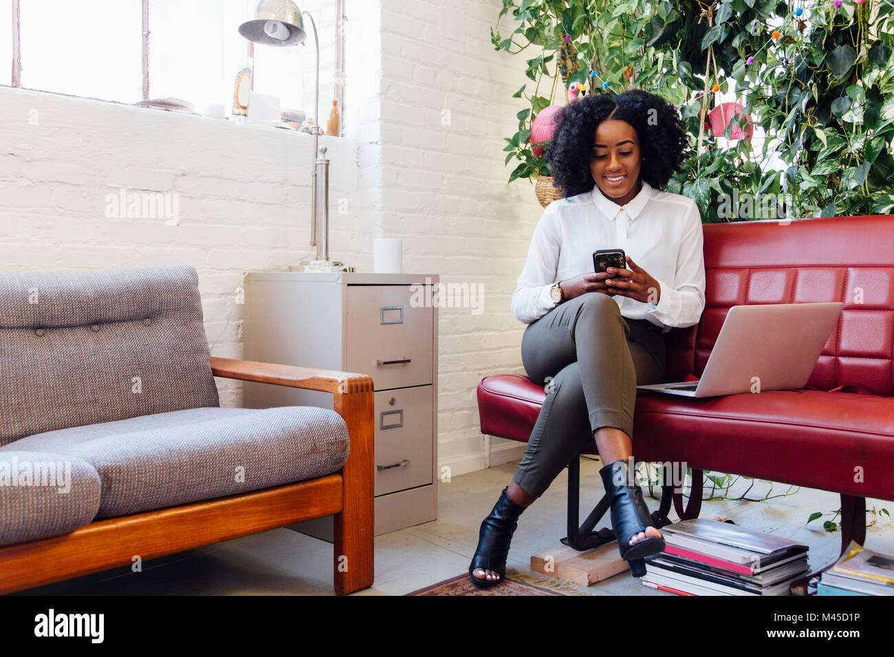 Woman in office sitting on sofa with laptop, texting on smartphone Stock Photo