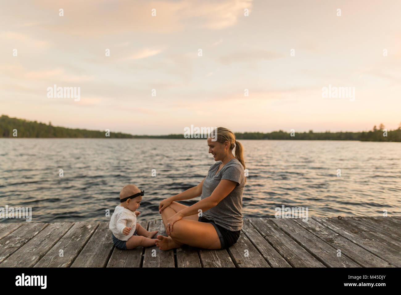Mother sitting cross legged on lake pier with baby daughter Stock Photo