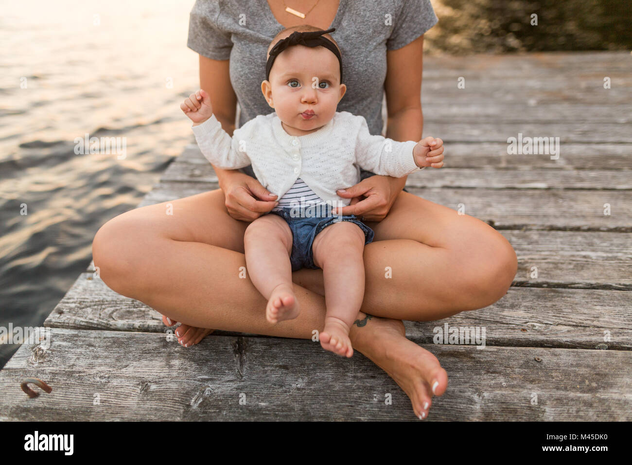 Mother sitting cross legged on lake pier with baby daughter on lap, cropped Stock Photo
