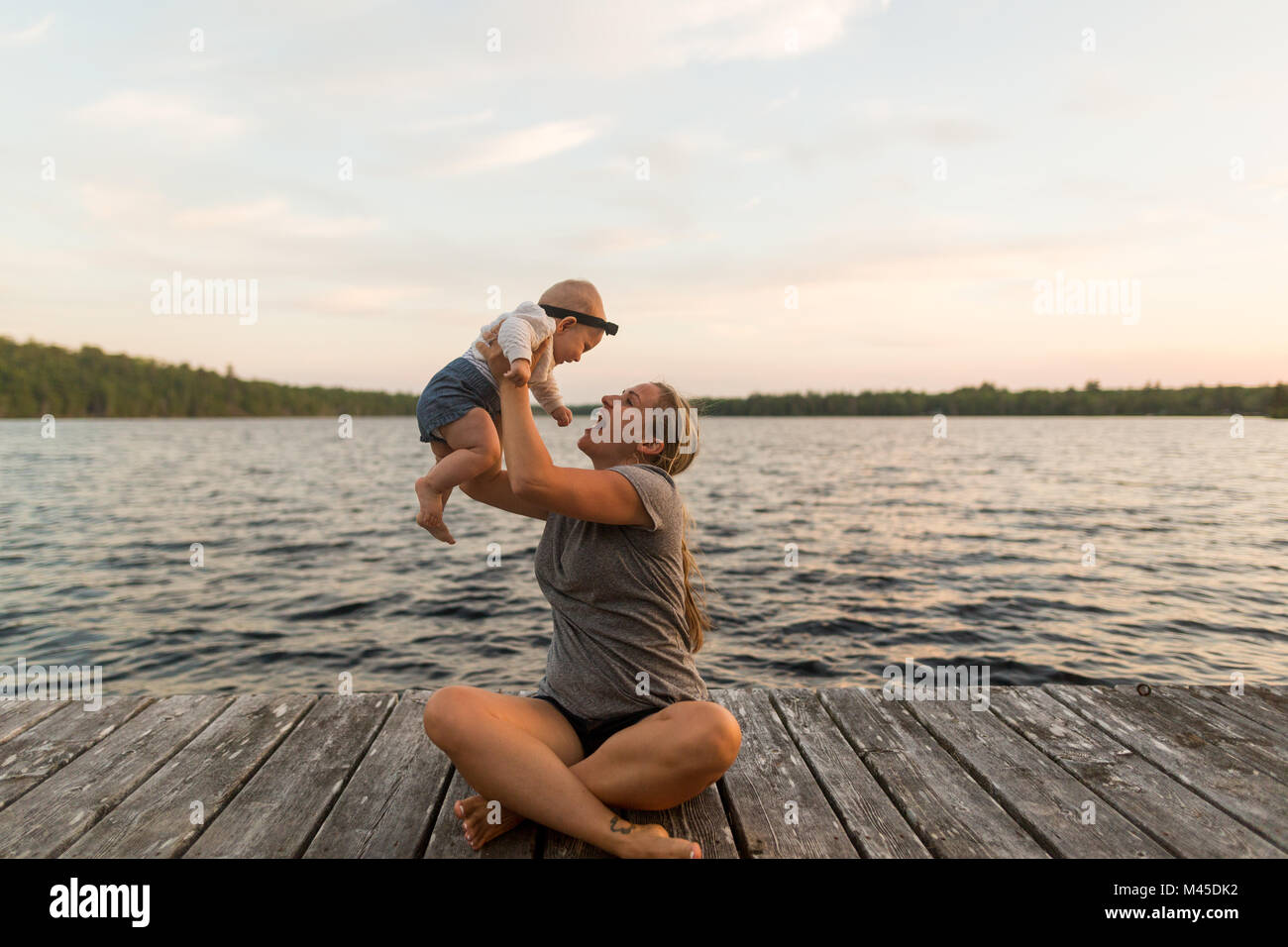 Mother sitting cross legged on lake pier holding up baby daughter Stock Photo