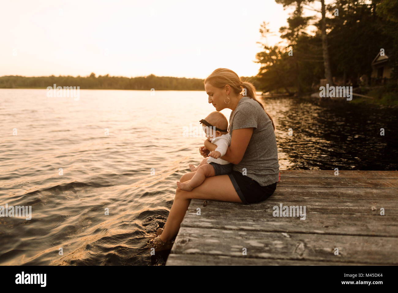 Mother sitting on pier with baby daughter looking down at  lake Stock Photo