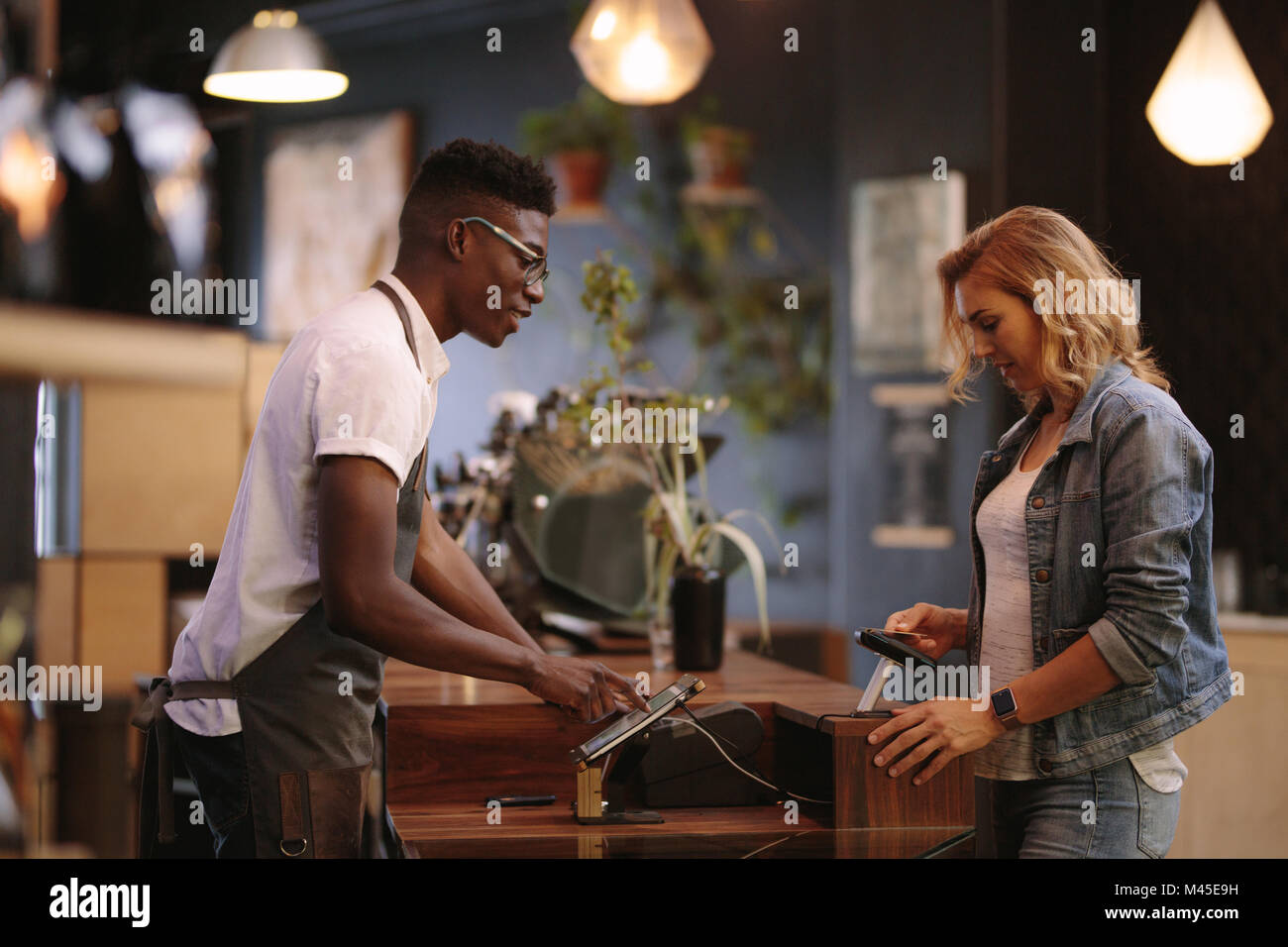 Coffee shop owner at the billing counter accepting payment from customer. Woman paying her coffee bills wirelessly using credit card. Stock Photo
