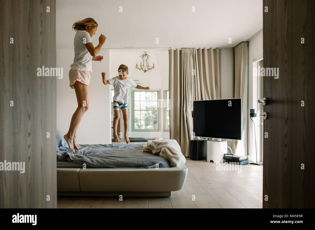 Young mother and her little son jumping on bed. Woman and little boy playing together and enjoying in bedroom. Stock Photo